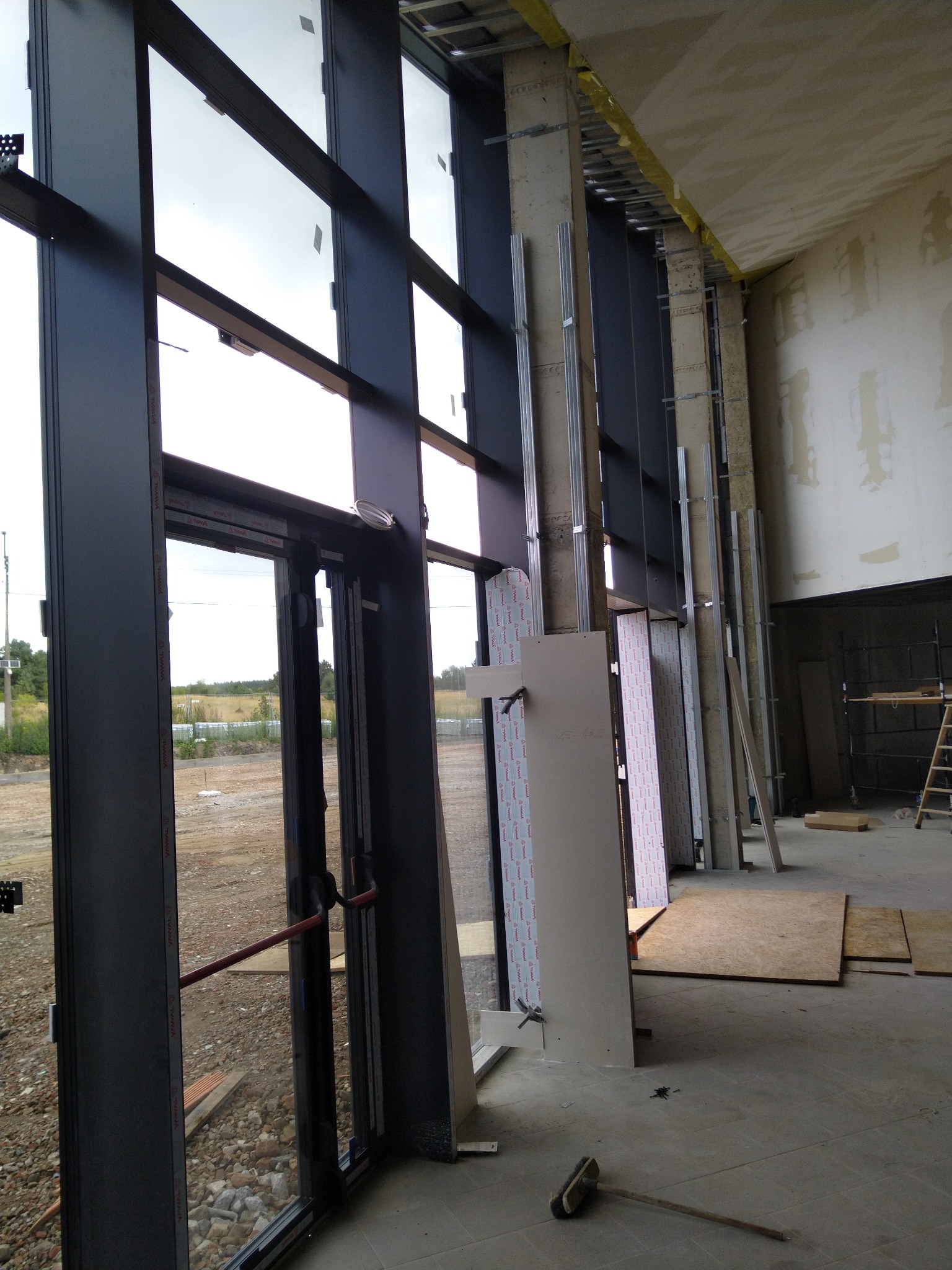 Wnętrze budynku w stanie surowym z zamontowanymi oknami i przygotowanymi elementami do montażu ścian działowych, widoczne profile metalowe i płyty, podłoże częściowo zabezpieczone płytami OSB.