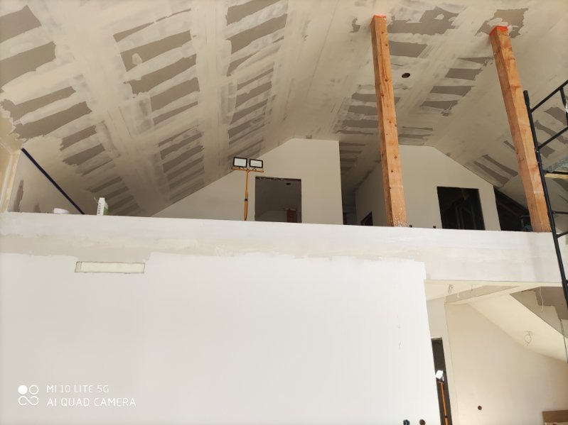 Skośny sufit poddasza w trakcie wykańczania płytami. Widoczne drewniane podpory i lampa budowlana. Na ścianach widoczne ślady szpachlowania.