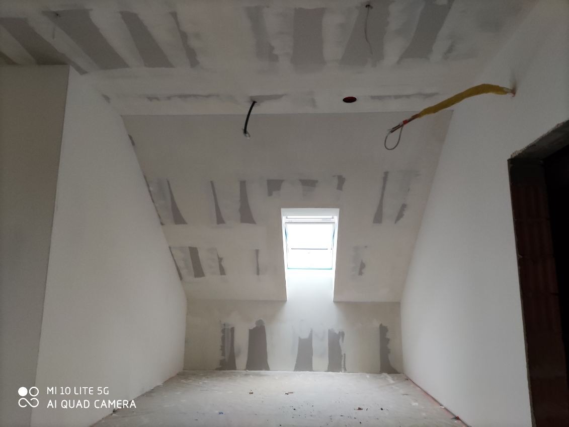 Poddasze z oknem dachowym w trakcie wykańczania: szpachlowane ściany i sufit przygotowane do malowania. Widoczne kable wystające ze ściany.