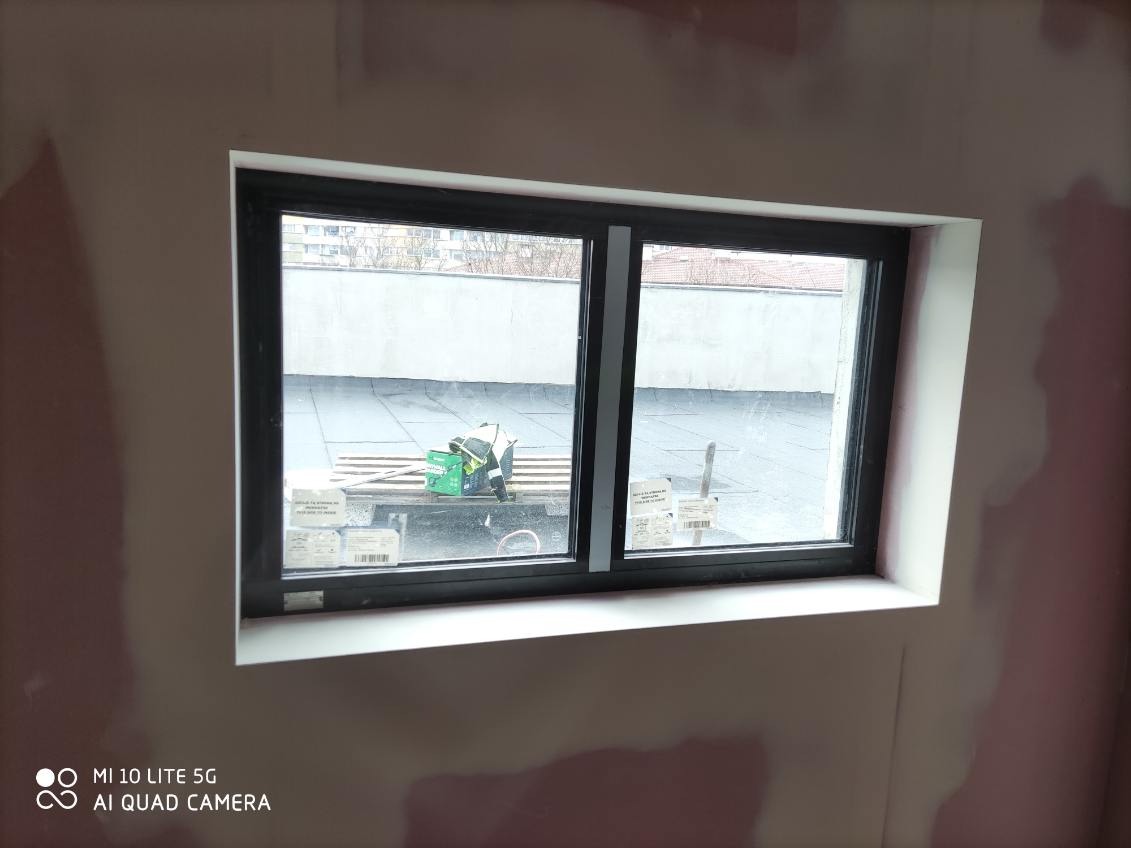 Nowe okno z czarną ramą zamontowane w ścianie, przed dalszym wykończeniem. Na zewnątrz widoczny pracownik na dachu. Ściana w trakcie szpachlowania.