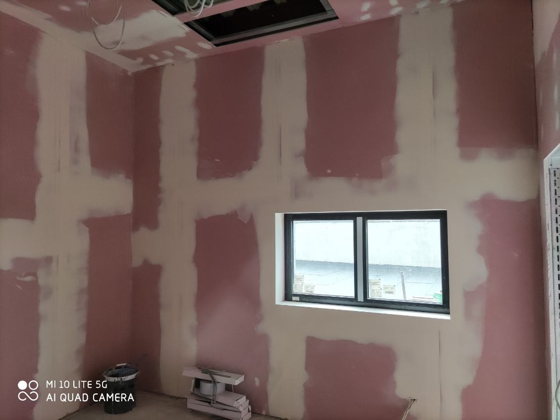 Wnętrze pokoju w trakcie prac wykończeniowych: różowe płyty gipsowe, białe łączenia, okno z czarną ramą, widoczny fragment sufitu podwieszanego i narzędzia.