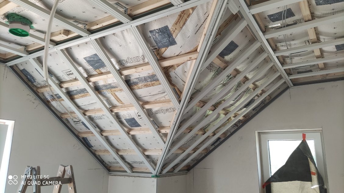 Skośny sufit poddasza w trakcie montażu: widoczna konstrukcja z profili, izolacja z folii aluminiowej. Okno z prawej strony, drabina w lewym dolnym rogu.