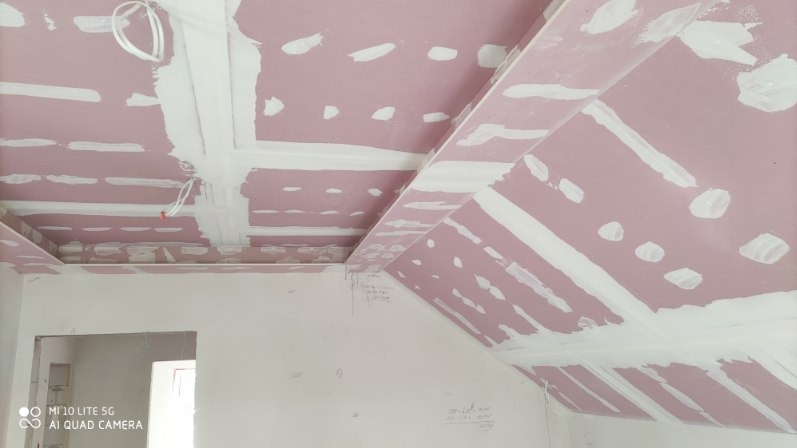 Wykończony sufit z płyt kartonowo-gipsowych, z widocznymi łączeniami szpachlowanymi na różowej płycie. Widoczne kable i fragment ściany z zapiskami.