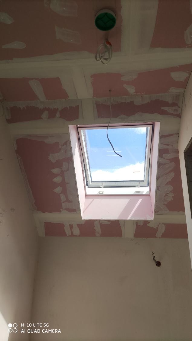 Montaż płyty KG na poddaszu z oknem dachowym, widoczne łączenia i instalacje elektryczne. Świeże łączenia płyt gipsowych, widok na błękitne niebo.