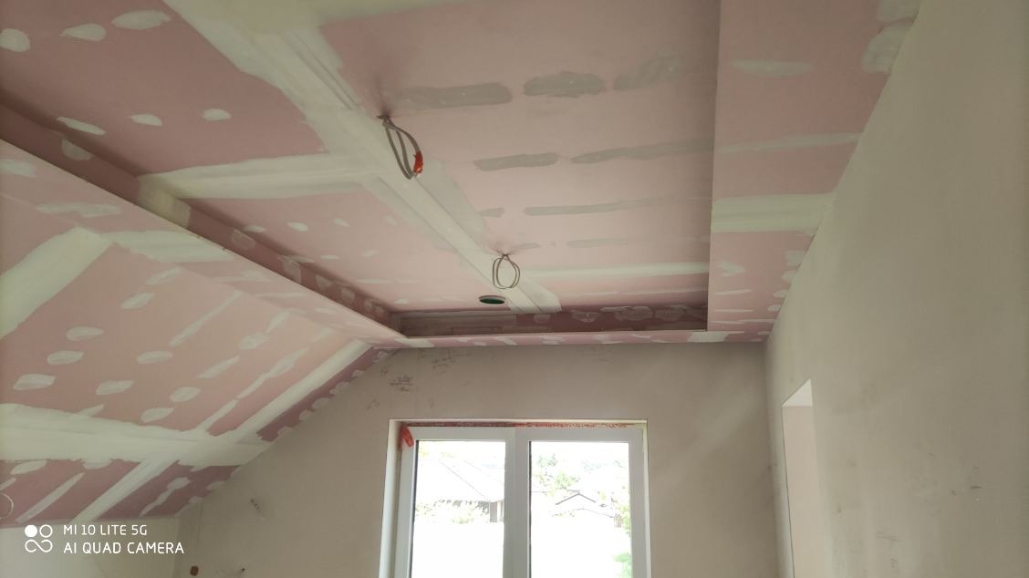 Sufit z płyt kartonowo-gipsowych w trakcie szpachlowania, widoczne kable oświetleniowe i okno. Prace wykończeniowe w nowym domu.