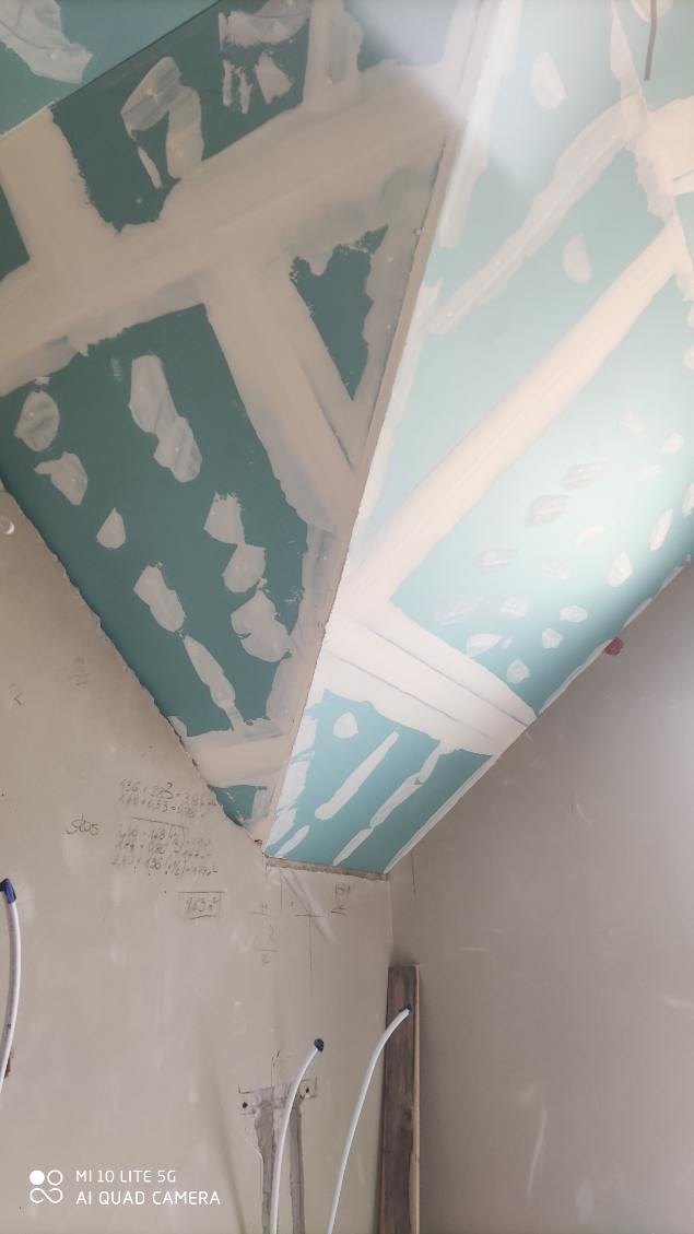 Montaż sufitu z płyty karton-gips w trakcie szpachlowania, widoczne połączenia płyt. Na ścianach odręczne notatki i wystające rurki instalacji.