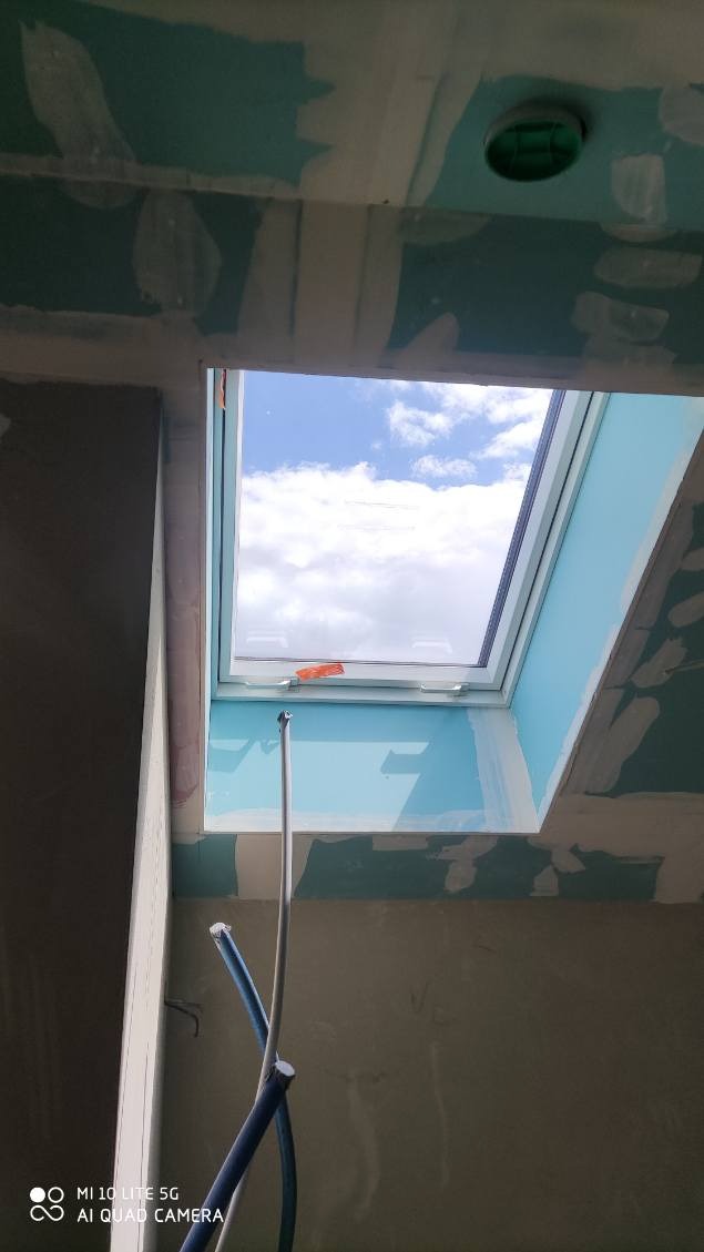 Montaż okna dachowego w suficie z płyty gipsowej. Widoczne niebieskie niebo i chmury przez okno. Prace wykończeniowe w trakcie.