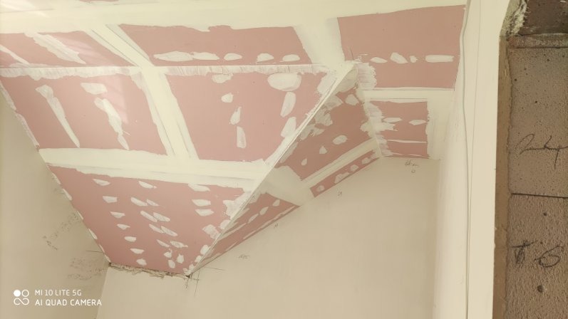 Skośny sufit z płyt kartonowo-gipsowych w trakcie szpachlowania, widoczne łączenia i różowa powierzchnia płyt. Fragment ściany z surowej cegły w tle.