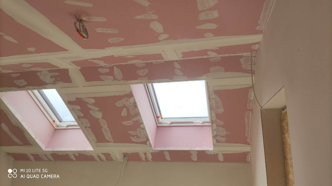 Montaż płyt karton-gips na poddaszu z oknami dachowymi, widoczne łączenia i szpachlowanie, surowy stan wykończenia wnętrza.