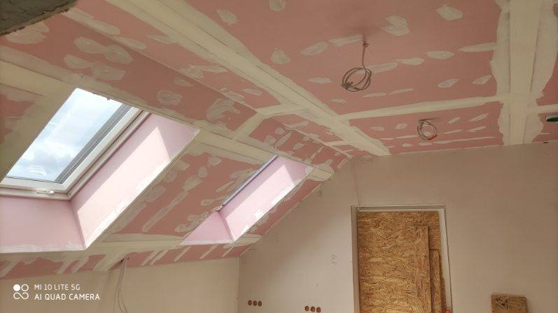 Montaż płyt karton-gips na poddaszu z oknami dachowymi. Widoczne łączenia płyt i wystające kable oświetleniowe. Ściany przygotowane do malowania.