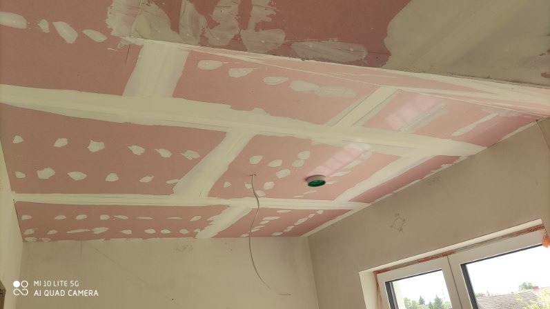 Różowe płyty z wyszpachlowanymi łączeniami na suficie podwieszanym, widok z dołu, surowy stan wykończenia, kabel oświetleniowy zwisa z sufitu.
