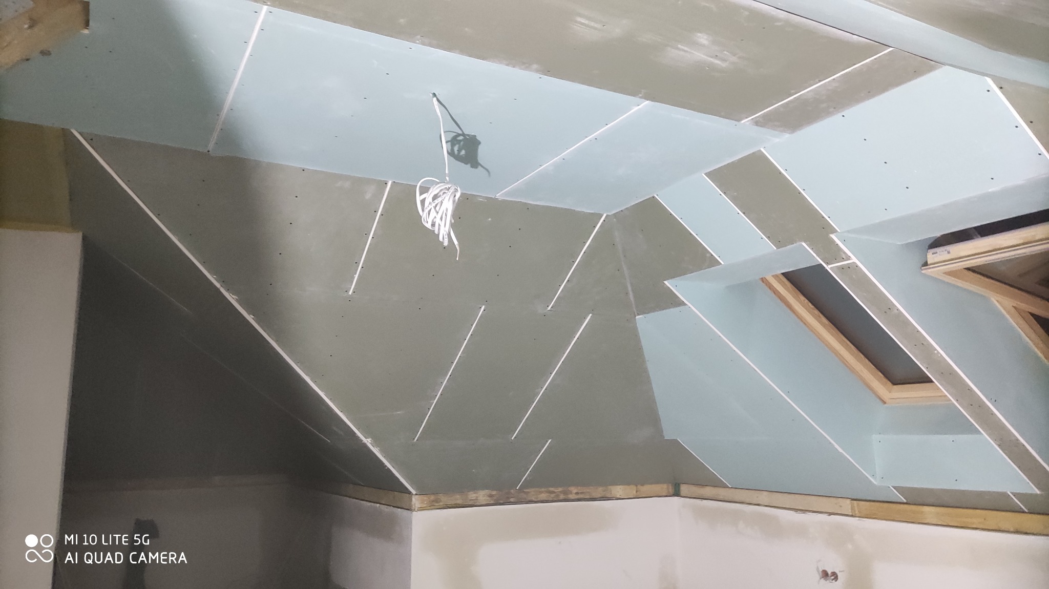 Skośny sufit poddasza z zamontowanymi płytami, widoczne kable. Prace wykończeniowe w Bielicach Nowych. Nietypowy układ płyt przy oknie dachowym.