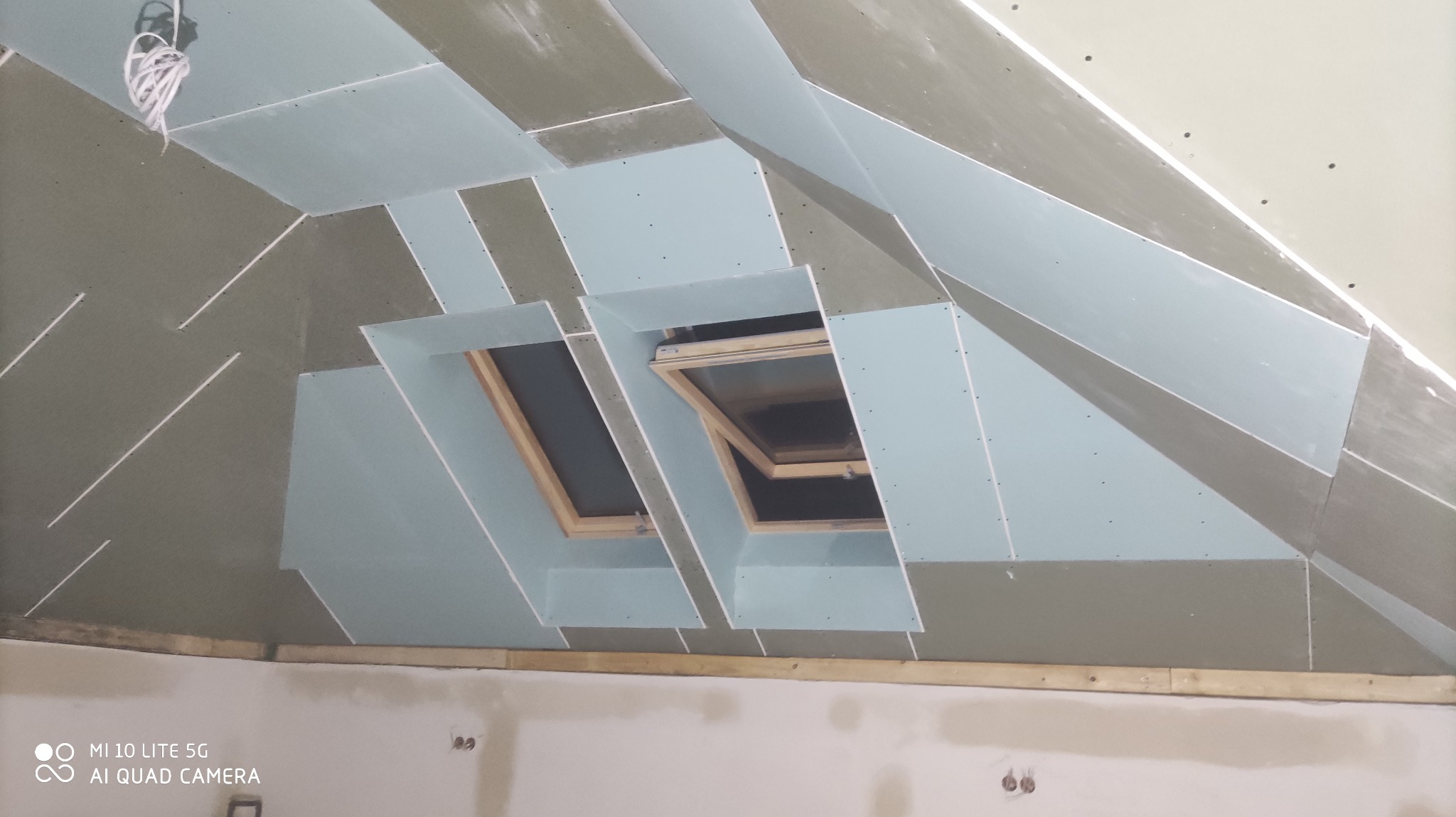 Skomplikowana konstrukcja sufitu poddasza z oknami dachowymi, częściowo pokryta płytami gipsowo-kartonowymi, widoczne łączenia i szpachlowanie.