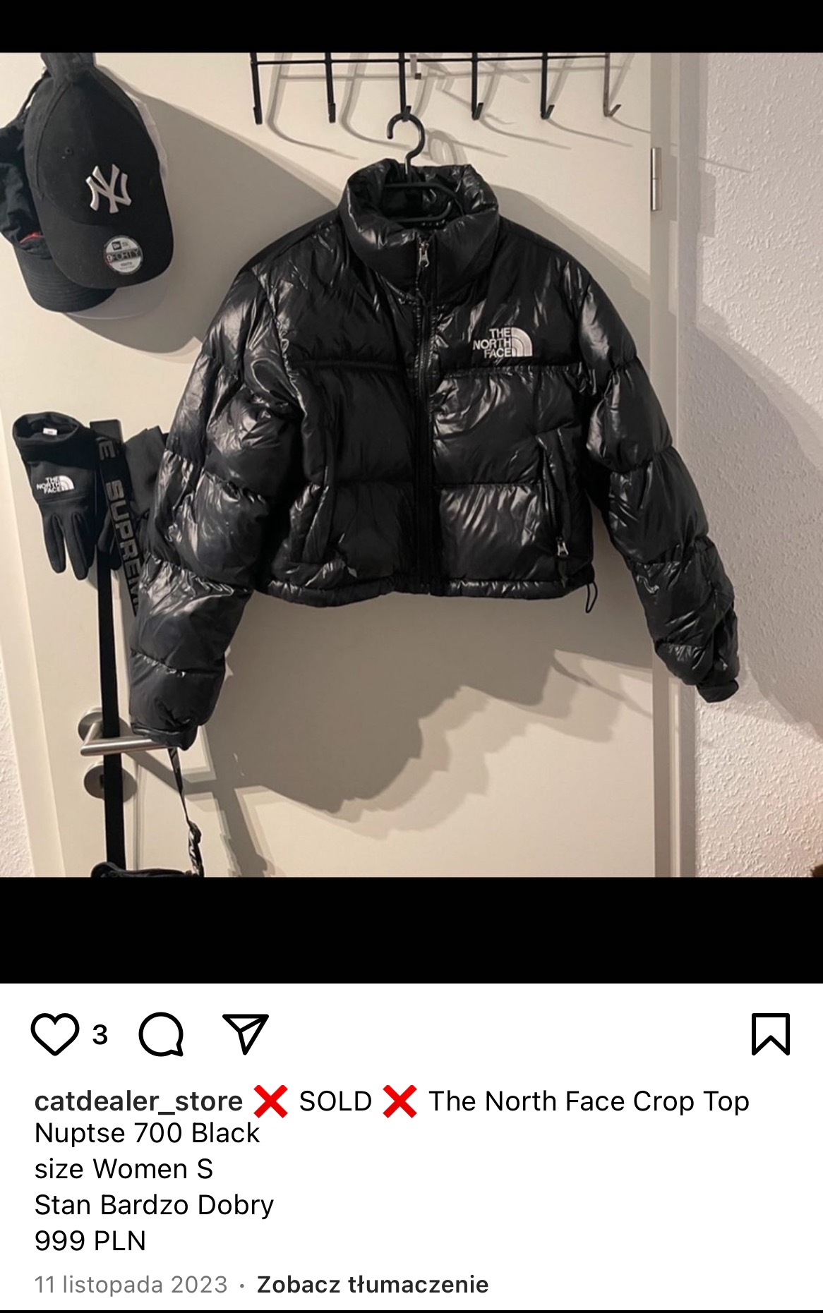 Czarna, krótka kurtka puchowa The North Face Nuptse 700 damska w rozmiarze S, wisząca na wieszaku na tle białych drzwi, obok czapka New York Yankees i czarne rękawice The North Face Supreme.