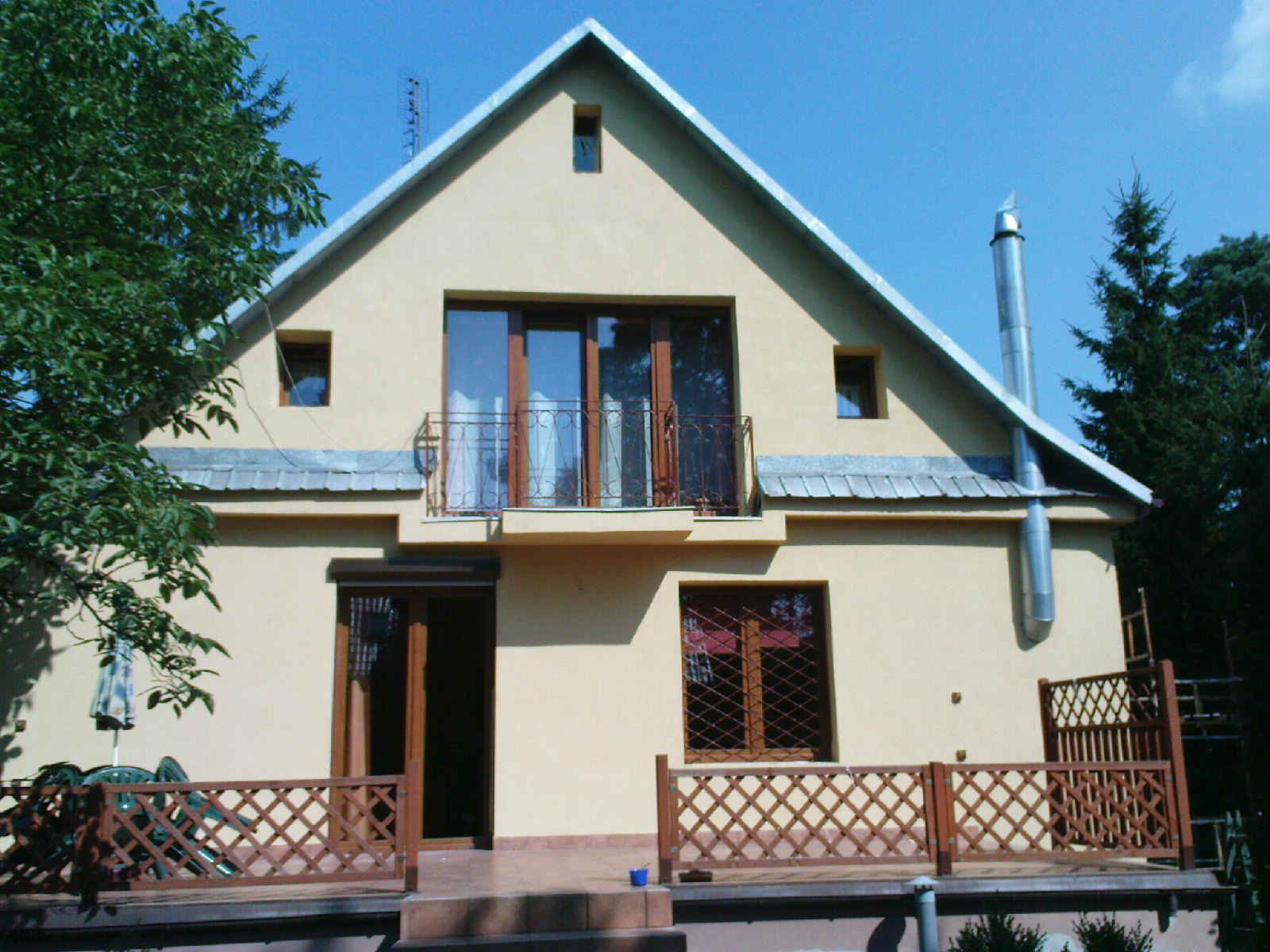 Żółta elewacja domu z balkonem i brązową stolarką okienną, widoczny komin ze stali nierdzewnej, ujęcie z poziomu gruntu.