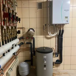 Zestaw urządzeń grzewczych w kotłowni: pompa ciepła Mitsubishi Ecodan, zasobnik Hajdu, liczne rury miedziane z zaworami i manometrami, oraz szare naczynie przeponowe, całość na tle płytek...