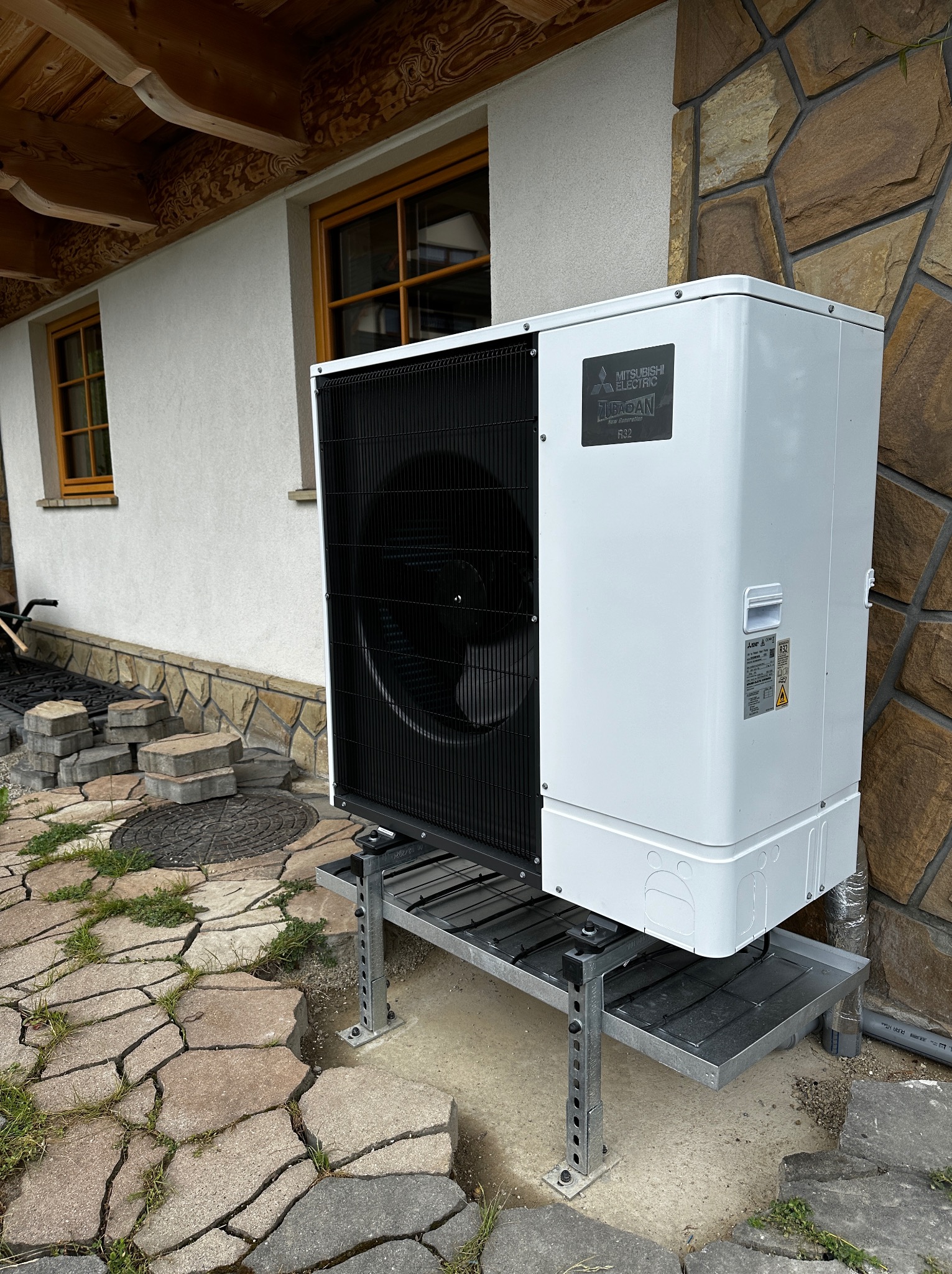 Zewnętrzna jednostka pompy ciepła Mitsubishi Electric Zubadan R32 na metalowej konstrukcji wsporczej, zamontowana przy ścianie budynku z elementami kamiennej elewacji i drewnianym wykończeniem...