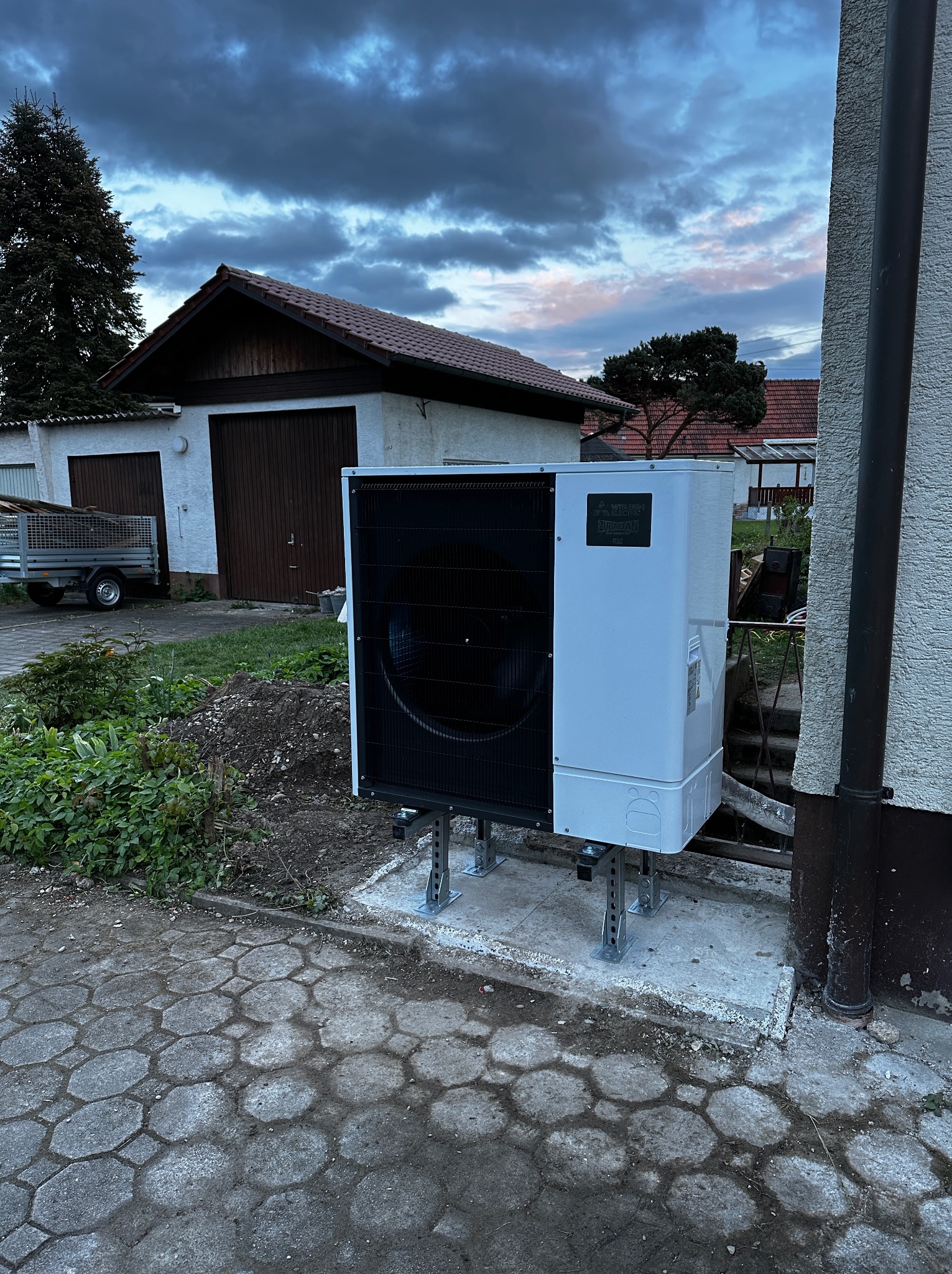 Biała jednostka zewnętrzna z czarnym wentylatorem, zamontowana na metalowych wspornikach na betonowej podstawie, na tle garażu i pochmurnego nieba o zmierzchu.