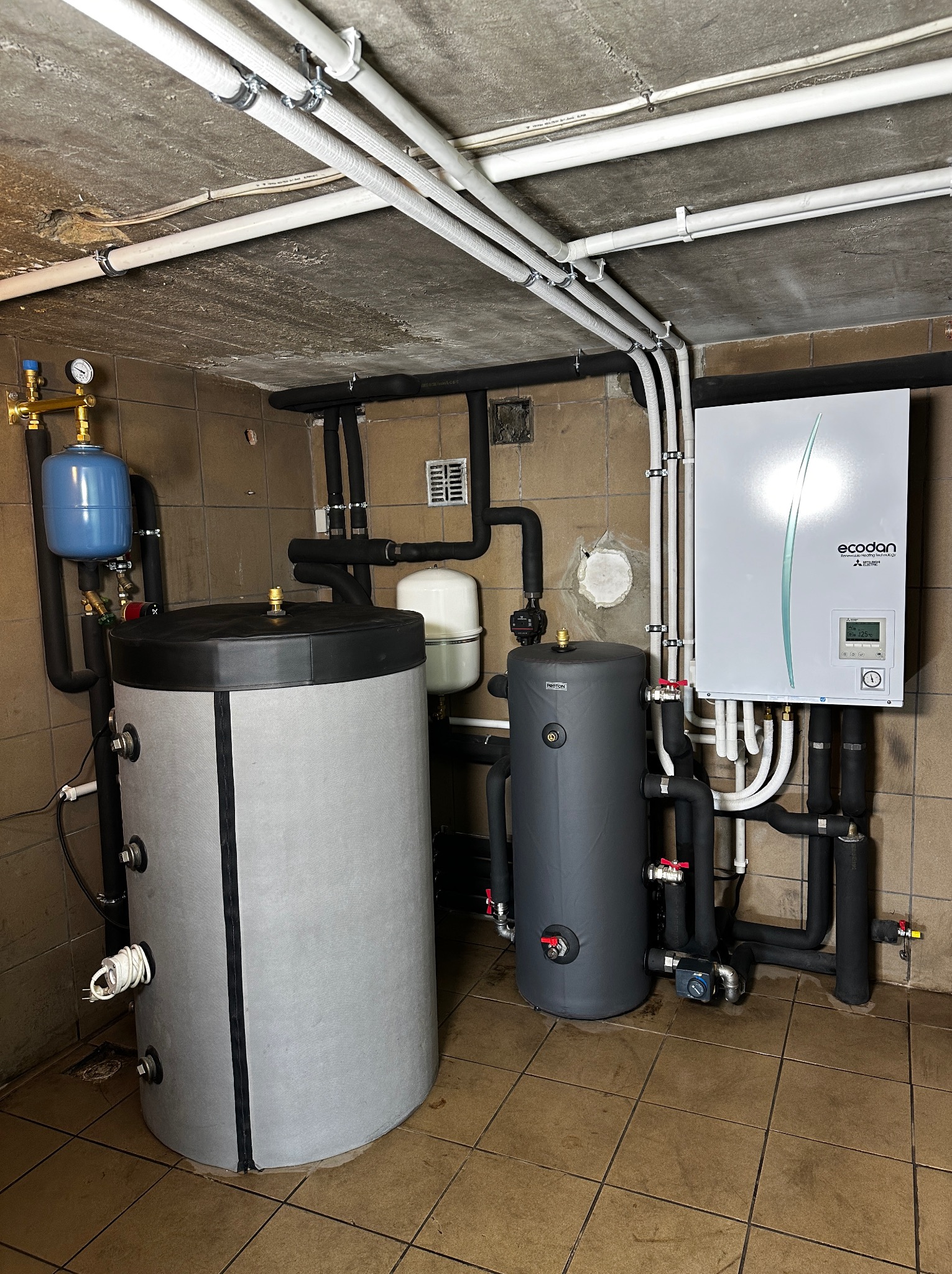 Instalacja w kotłowni: duży szary zbiornik, niebieski zbiornik wyrównawczy, biały zbiornik i biała jednostka Ecodan z podłączeniami rur w piwnicy z brązowymi płytkami podłogowymi.