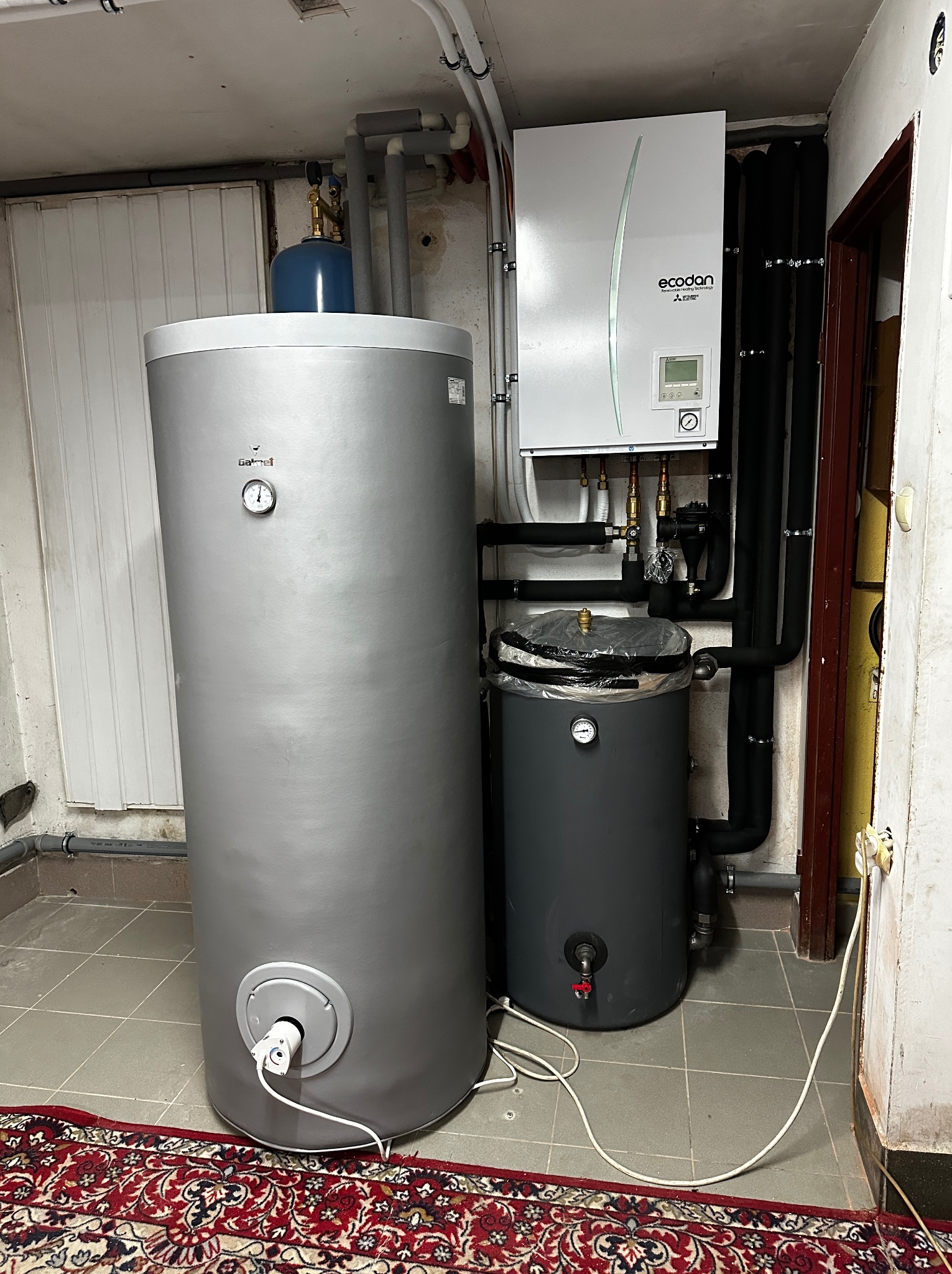 Szary zasobnik ciepłej wody użytkowej marki Galmet z podłączonym przewodem elektrycznym, obok mniejszy, ciemnoszary zbiornik z zaworem, na tle ściany z rurami i jednostką wewnętrzną pompy ciepła...