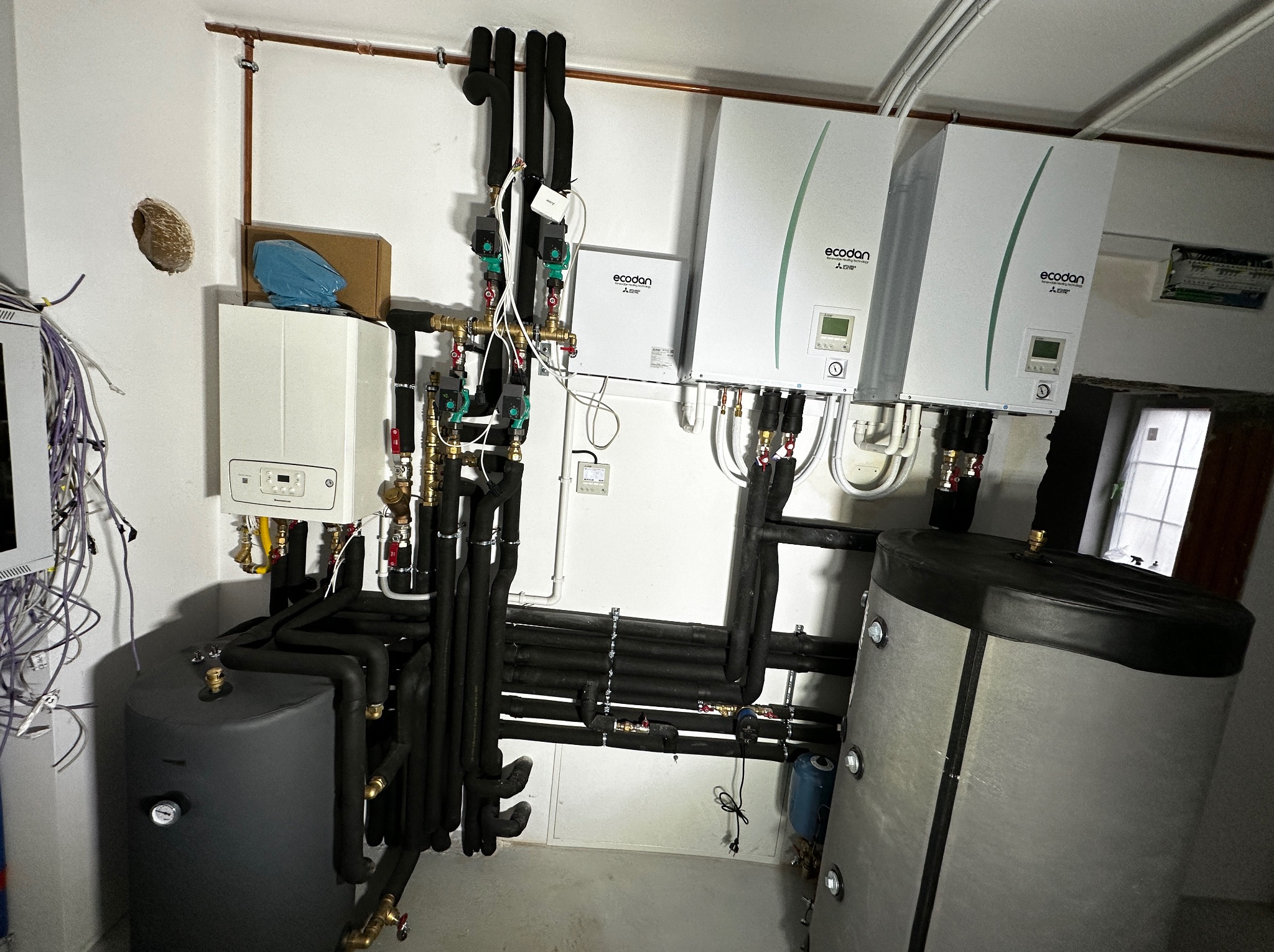 Instalacja pompy ciepła Ecodan Mitsubishi z zasobnikiem, widoczne liczne rury, zawory i okablowanie, zainstalowane w pomieszczeniu technicznym.