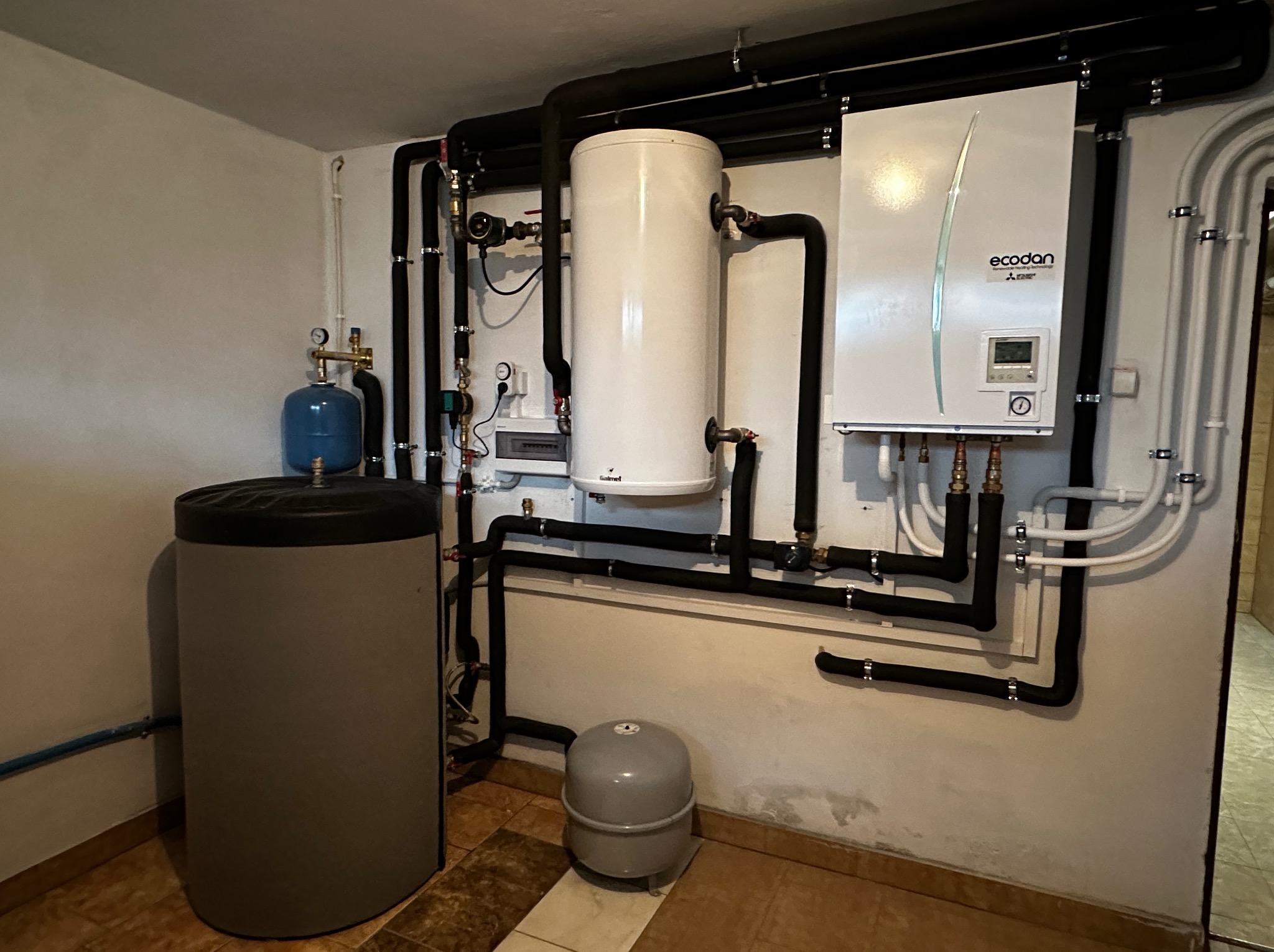 Zestaw urządzeń grzewczych w kotłowni: zbiornik buforowy, zasobnik ciepłej wody i jednostka zewnętrzna z czarnymi rurami w izolacji.