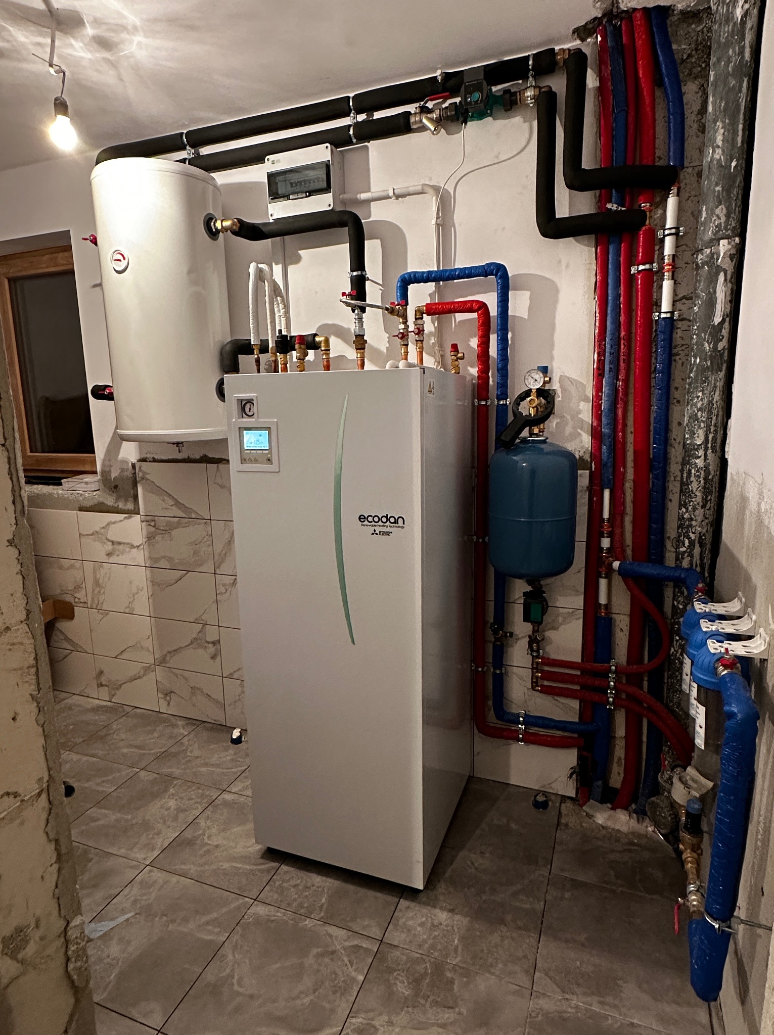 Nowoczesna instalacja grzewcza z zasobnikiem i rurami w kolorach niebieskim i czerwonym, widoczny manometr i zawory, tło stanowią płytki imitujące marmur.