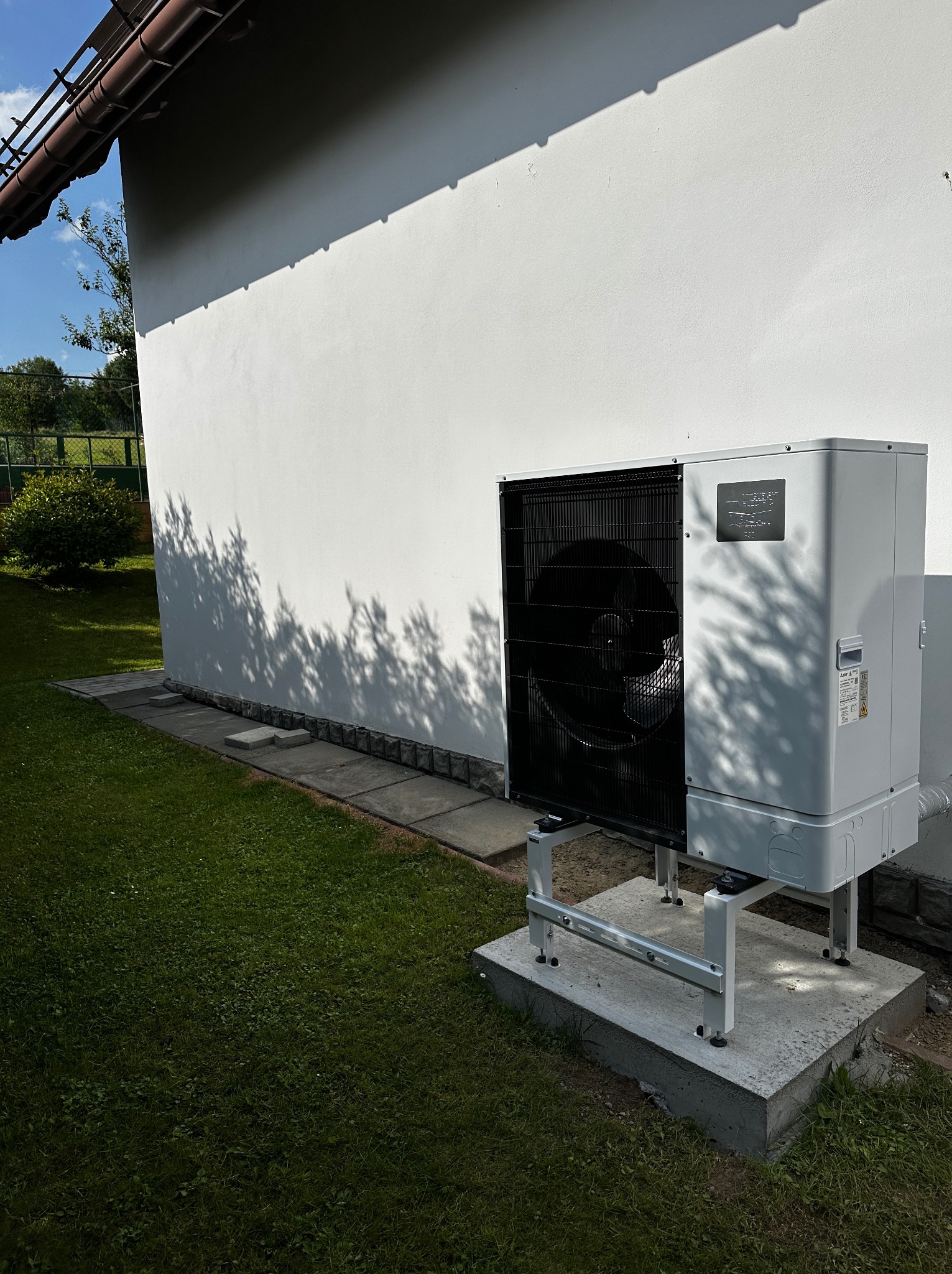 Zewnętrzna jednostka pompy ciepła Mitsubishi zamontowana na betonowej podstawie przy ścianie budynku, z widocznymi cieniami drzew na elewacji.