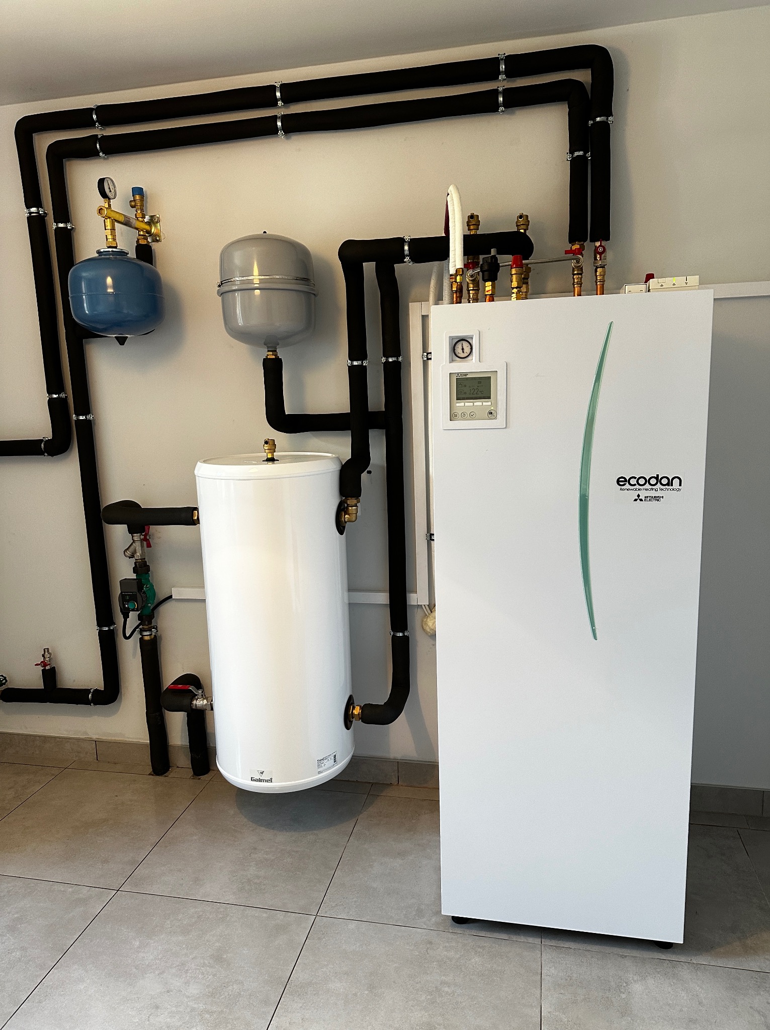 Biała pompa ciepła Ecodan Mitsubishi Electric z podłączonymi rurami w izolacji, zbiornik wyrównawczy ciśnienia w kolorze niebieskim i szarym oraz biały zasobnik ciepłej wody użytkowej w kotłowni...