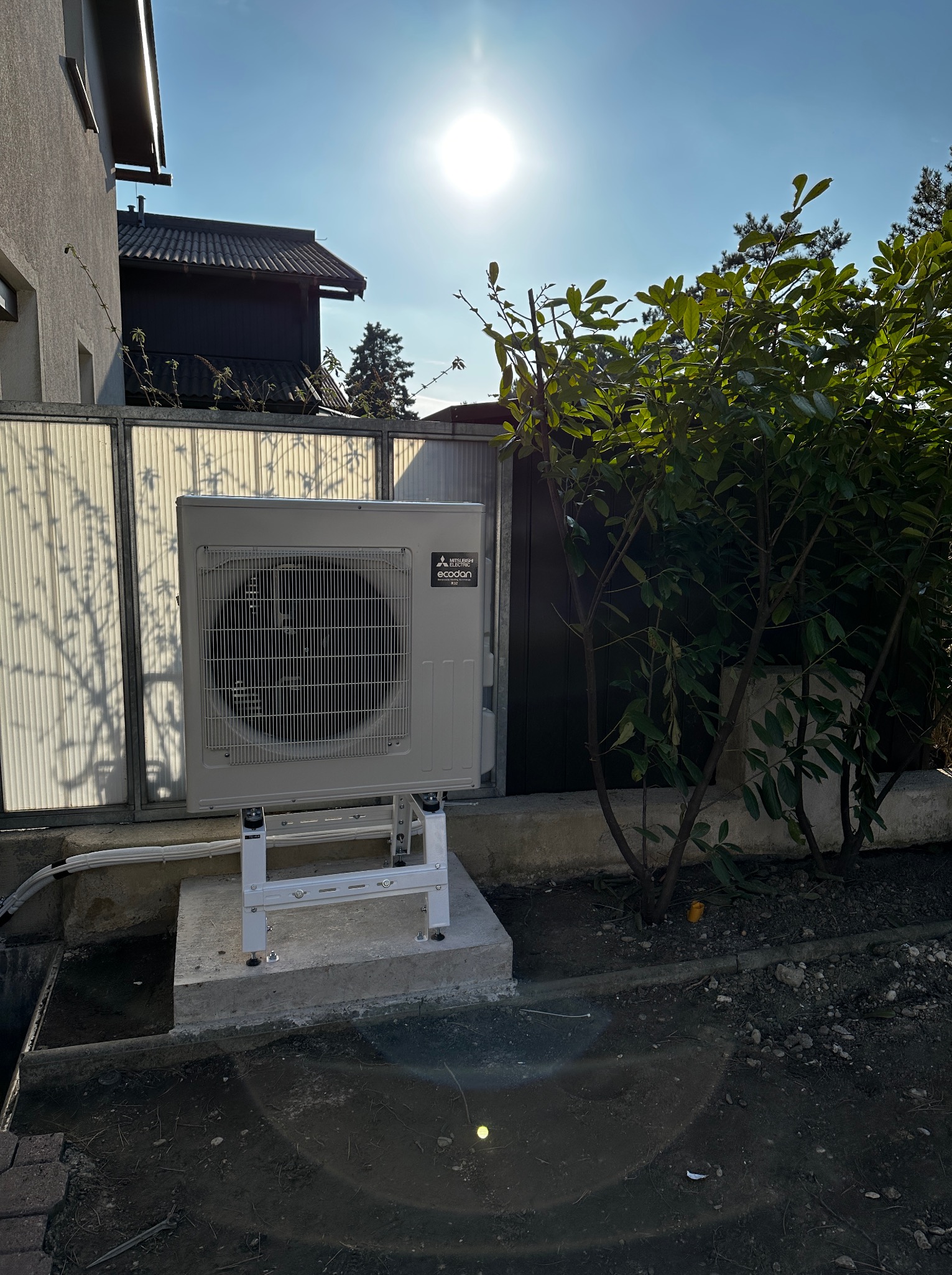 Zewnętrzna jednostka klimatyzacji Mitsubishi Ecodan zamontowana na betonowym fundamencie i metalowej konstrukcji, widoczna z boku na tle zieleni i części budynku w słoneczny dzień.