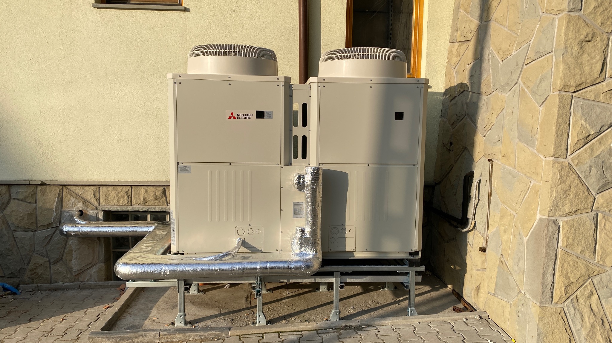 Dwie jednostki zewnętrzne pompy ciepła Mitsubishi Electric, z izolowanymi rurami, umieszczone na metalowej konstrukcji przy budynku o elewacji z kamienia łupanego.