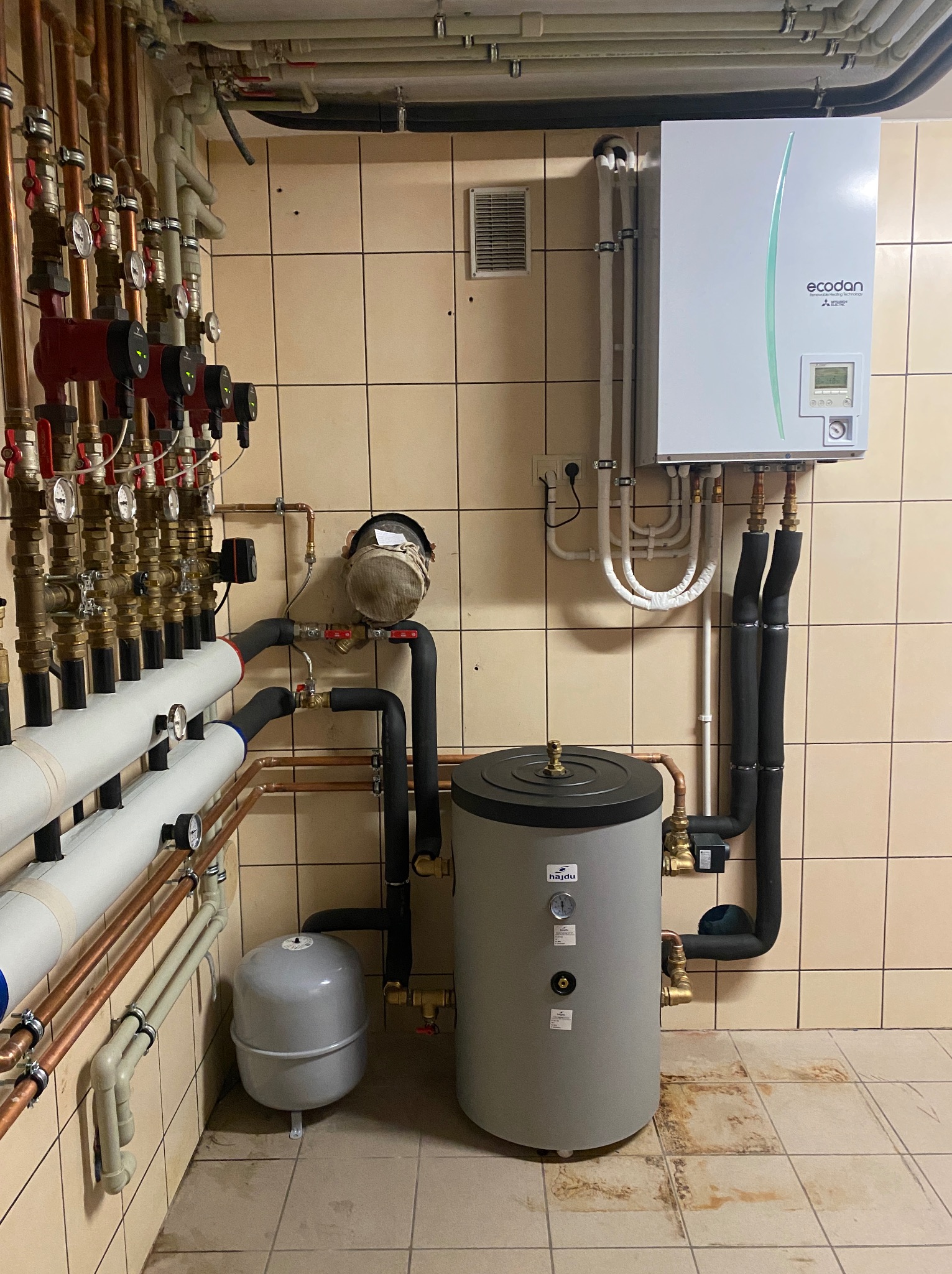 Kotłownia z zainstalowaną pompą ciepła Mitsubishi Ecodan, zasobnikiem Hajdu, licznymi rurami miedzianymi i białymi, manometrami oraz szarą przeponą na tle ceramicznych płytek.