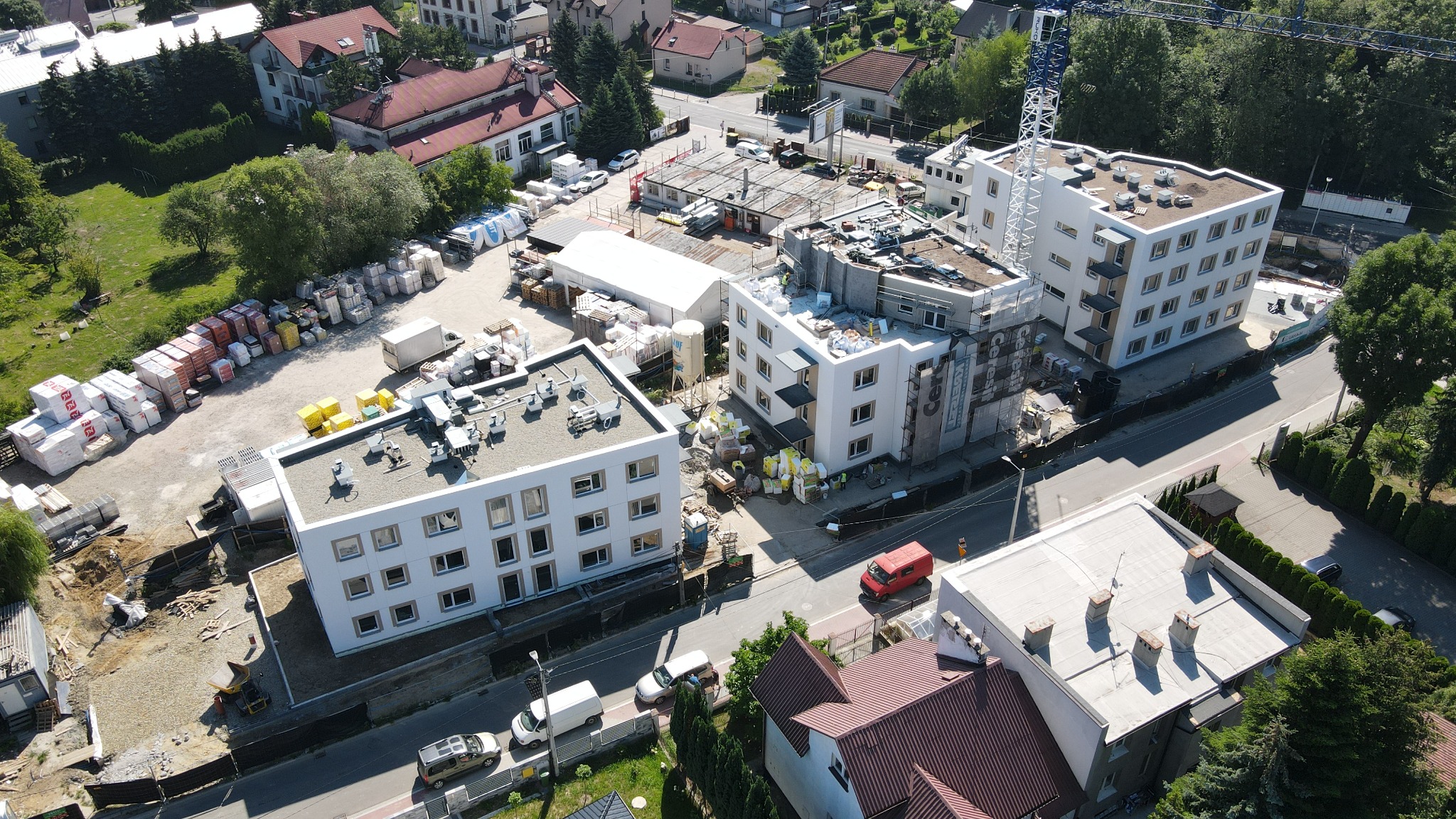 Widok z góry na plac budowy z kilkoma budynkami w trakcie wznoszenia, materiały budowlane składowane obok, żuraw budowlany w tle, czerwony samochód dostawczy na ulicy.