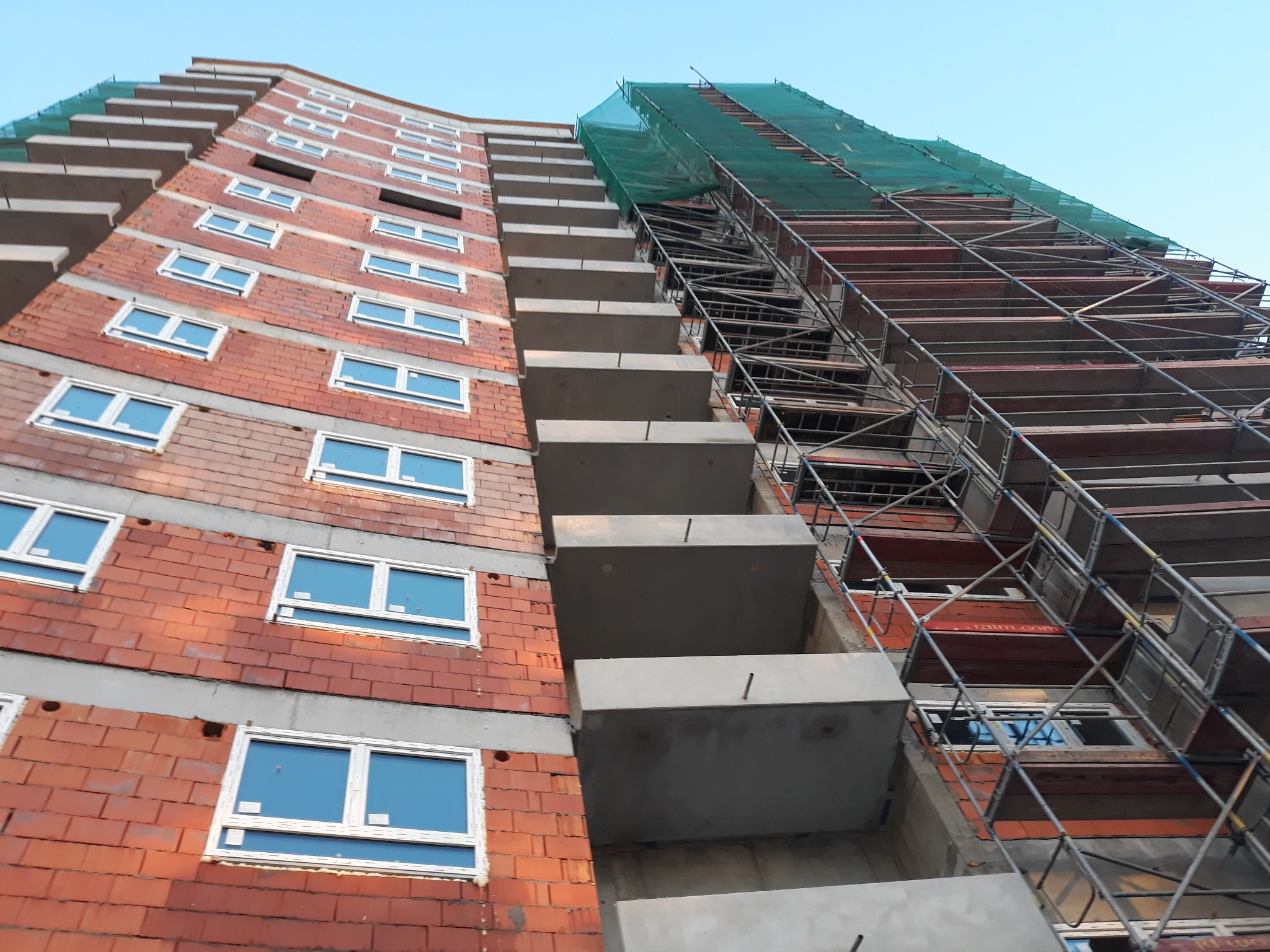 Wznoszenie wielopiętrowego budynku z cegły z widocznymi balkonami i rusztowaniami zabezpieczonymi siatką ochronną, ujęcie z dołu w słoneczny dzień.