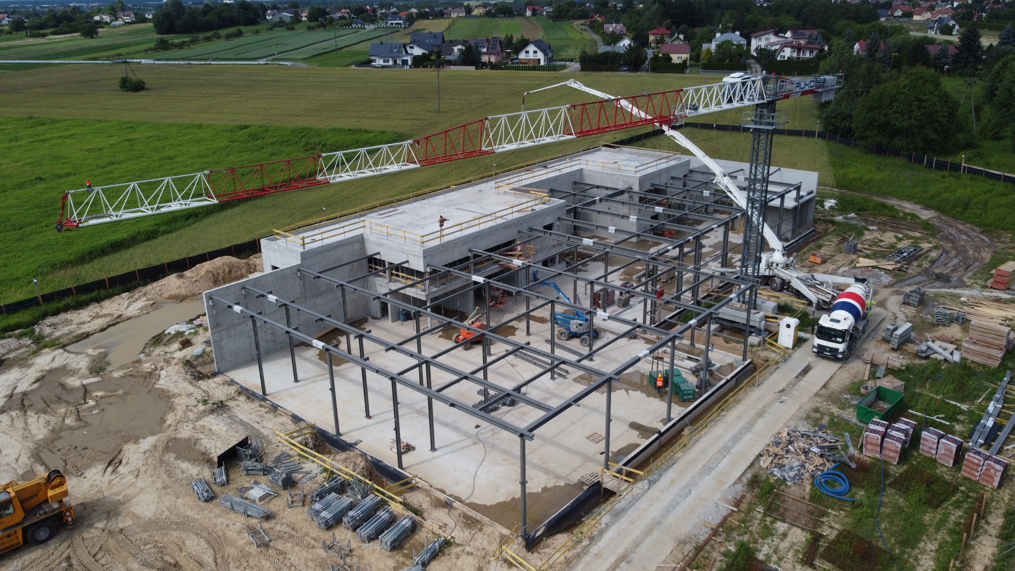 Widok z góry na plac budowy z betonową konstrukcją w trakcie wznoszenia, stalowym szkieletem, żurawiem budowlanym z czerwono-białym ramieniem i betoniarką w tle.