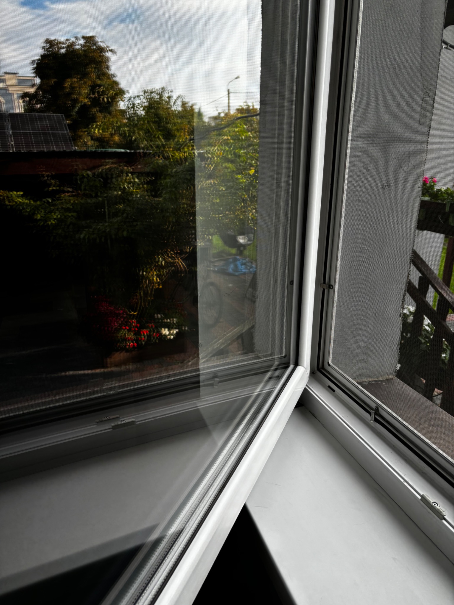 Otwarty narożny fragment okna z moskitierą, widok na zewnątrz, gdzie widać zieleń, fragment budynku z panelami słonecznymi i rower.