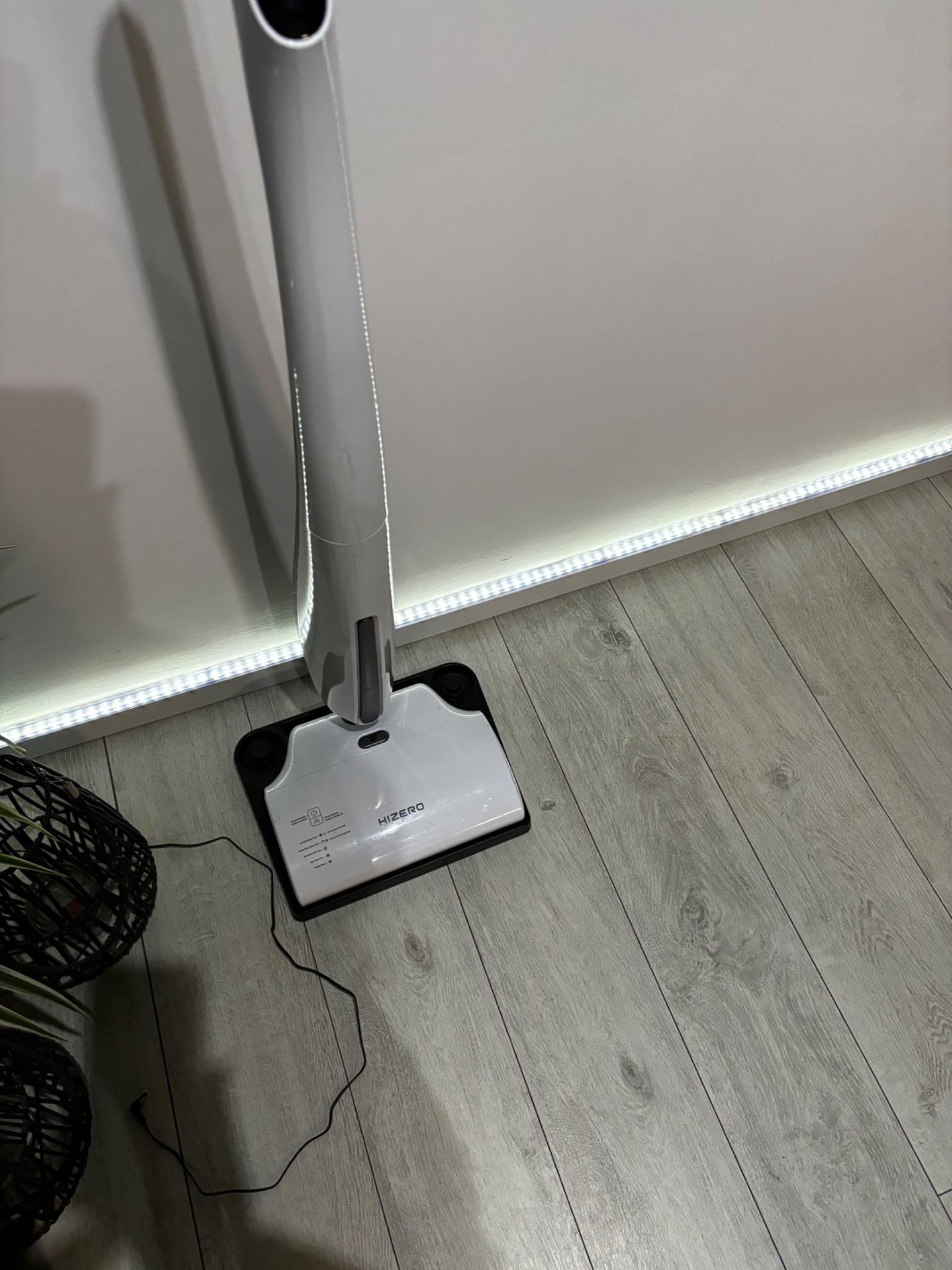 Bezprzewodowy mop elektryczny Hizero stojący na panelach podłogowych, z widocznym przewodem zasilającym i doniczką z rośliną w tle, oświetlony listwą LED.