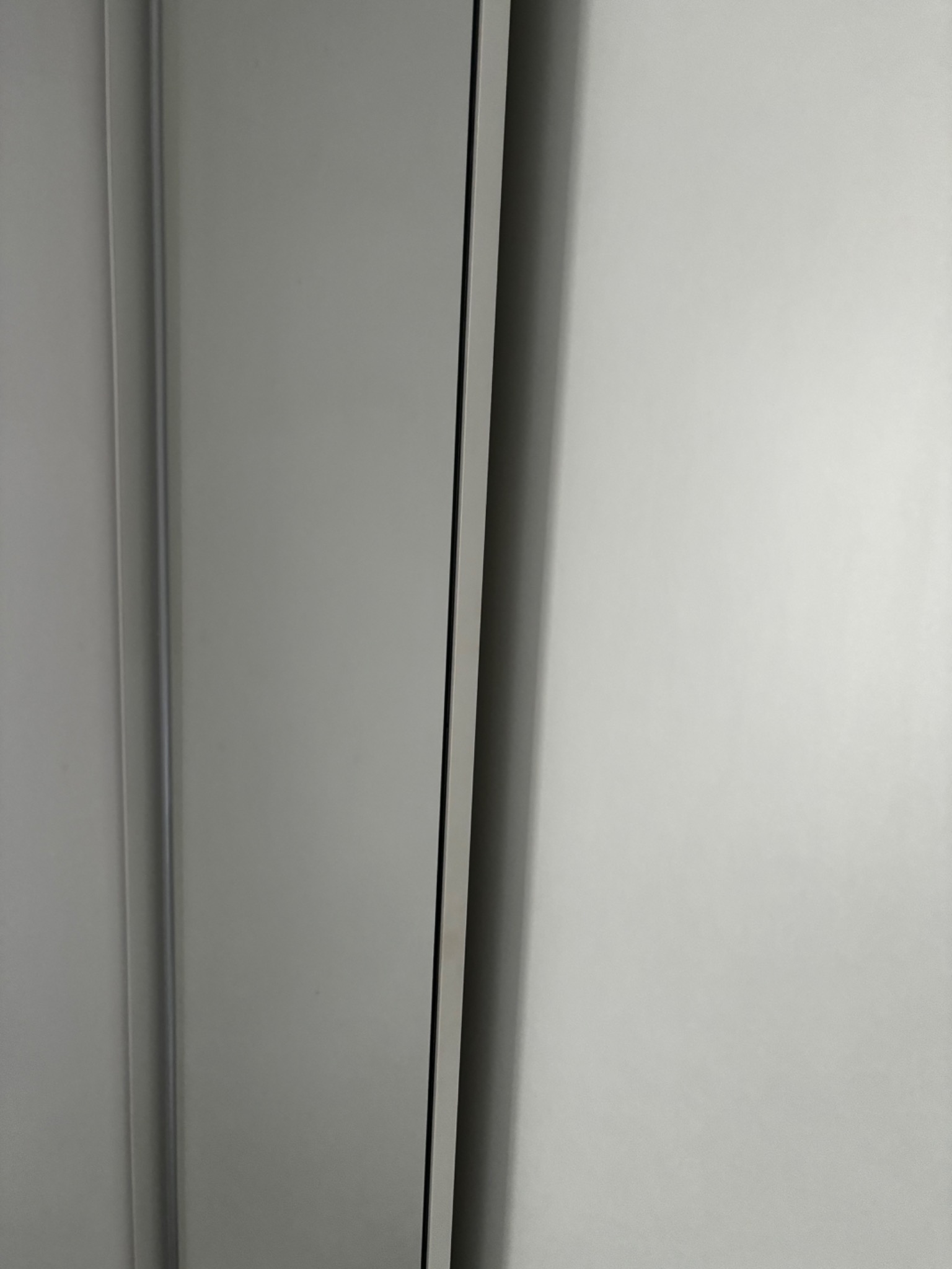 Gładka, jasnoszara powierzchnia szafy lub drzwi, z widoczną pionową krawędzią i delikatnym cieniem.