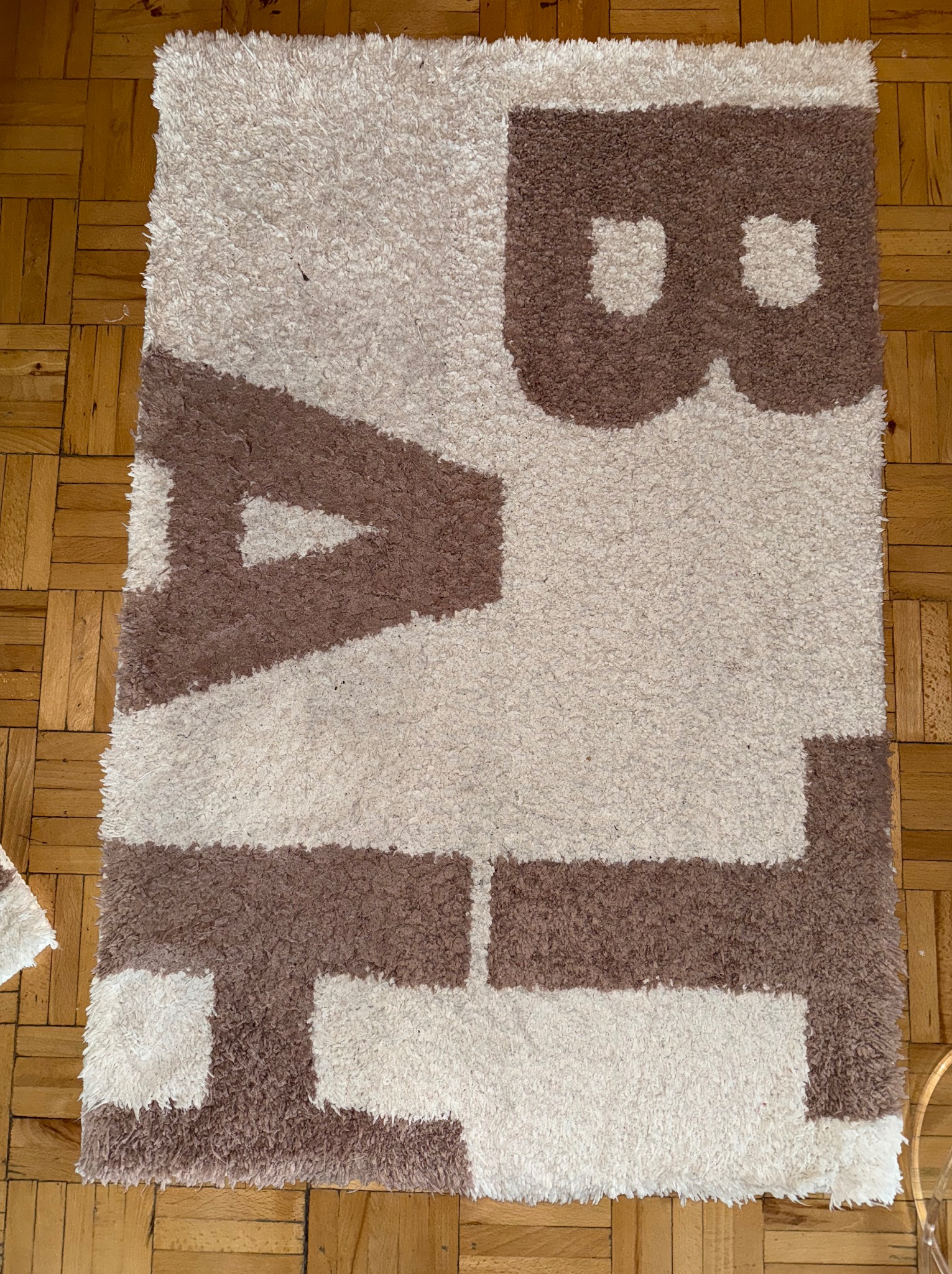 Beżowo-brązowy, włochaty dywan z abstrakcyjnym wzorem liter na parkiecie. Widok z góry, miękka tekstura, ciepłe kolory, domowa atmosfera.