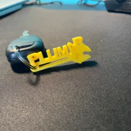 Żółty brelok z napisem 'Plumber' w kształcie klucza francuskiego, przymocowany do kluczyka samochodowego na ciemnym, teksturowanym podłożu.