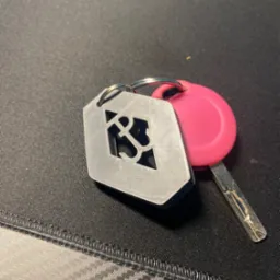 Klucz z r&oacute;żową obudową i metalowym brelokiem z wyciętym logo na czarnym tle z fakturą wł&oacute;kna węglowego.