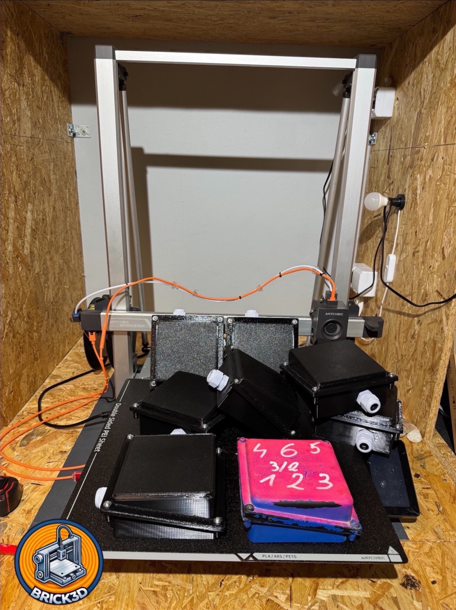 Drukarka 3D Anycubic Kobra 3 Max w drewnianej obudowie, drukująca czarne obudowy z tworzywa. Jedna z nich pomalowana na różowo-niebiesko z białymi numerami.