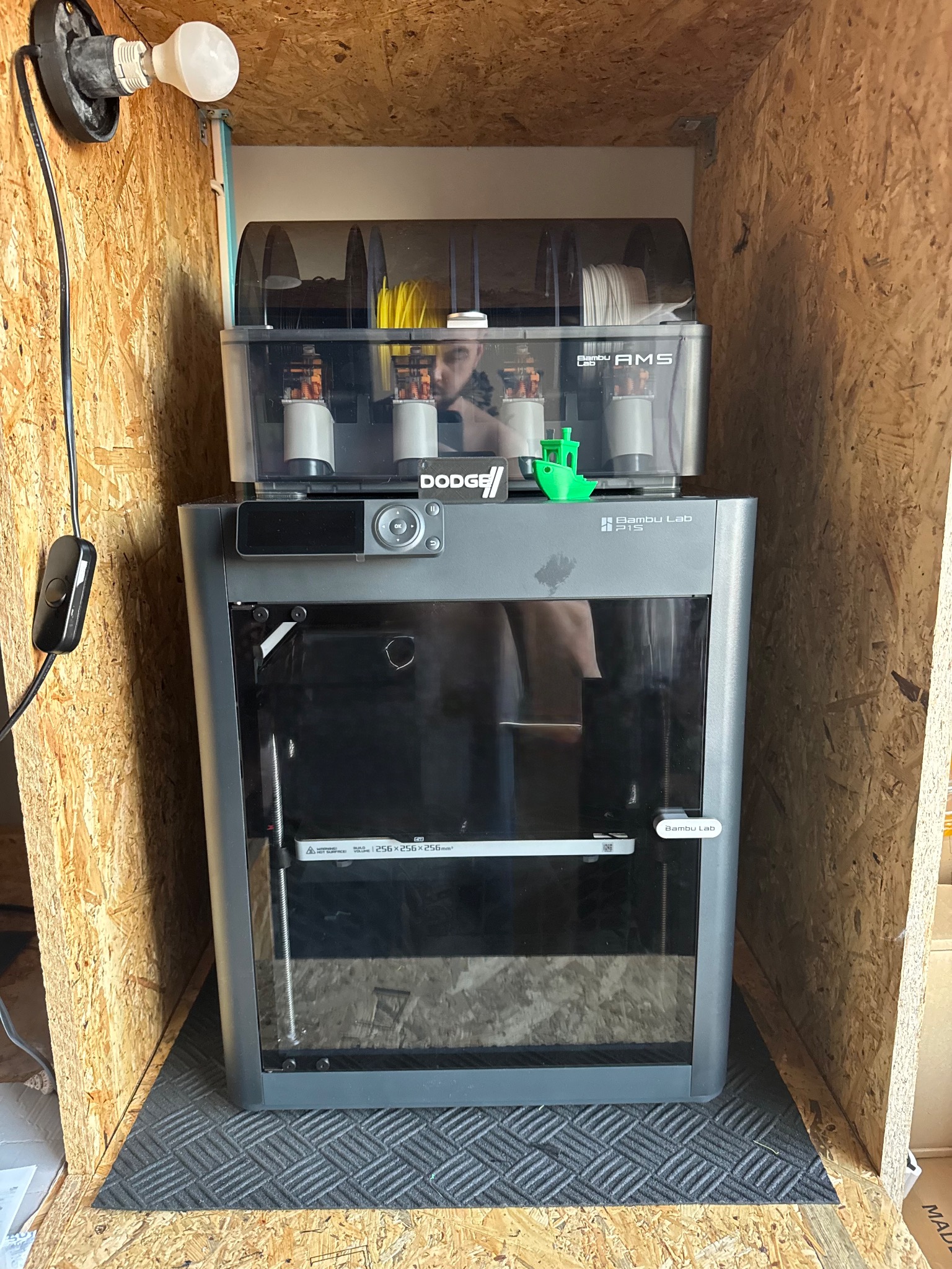 Drukarka 3D Bambu Lab P1S z systemem AMS w obudowie z płyty OSB, oświetlona żarówką. Na drukarce zielony model statku. Widoczne filamenty w podajniku.