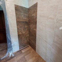 IMD-BUD - Wykończona łazienka z brązowo-beżowymi płytkami na ścianach i podłodze imitującej drewno; widoczne przyłącza wodne i odpływ liniowy, zabezpieczone folią drzwi.