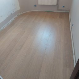 IMD-BUD - Ułożona podłoga z paneli imitujących drewno w jasnym odcieniu w pokoju z grzejnikiem na ścianie i przygotowaną ścianą do dalszych prac wykończeniowych.