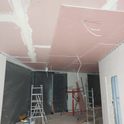 IMD-BUD - Montaż sufitu z płyt karton-gips: widoczne płyty w kolorze różowym, częściowo zaszpachlowane łączenia, wystające przewody elektryczne, drabiny rozstawione w pomieszczeniu.