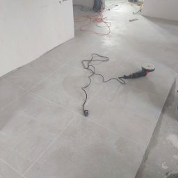 IMD-BUD - Świeżo położone, jasnoszare płytki podłogowe, częściowo mokre, z widoczną polerką do płytek, kablem zasilającym, ołówkiem i wiadrem w rogu pomieszczenia w trakcie prac wykończeniowych.