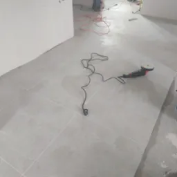 Świeżo położone, jasnoszare płytki podłogowe, częściowo mokre, z widoczną polerką do płytek, kablem zasilającym, ołówkiem i wiadrem w rogu pomieszczenia w trakcie prac wykończeniowych.