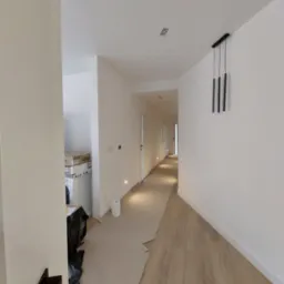 Korytarz w trakcie prac wykończeniowych: podłoga częściowo zabezpieczona kartonem, białe ściany z zamontowanym oświetleniem punktowym i wiszącą lampą, widoczne materiały budowlane w tle.