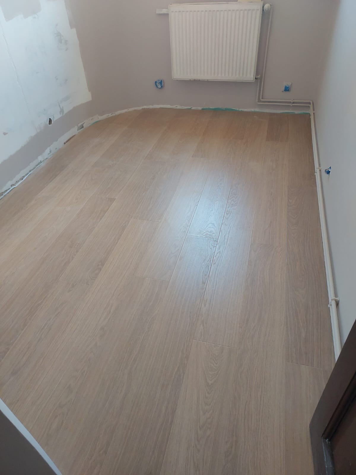 Ułożona podłoga z paneli imitujących drewno w jasnym odcieniu w pokoju z grzejnikiem na ścianie i przygotowaną ścianą do dalszych prac wykończeniowych.